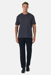 Námořnická modrá trička s krátkým rukávem a kulatým výstřihem, v kombinaci s černými kalhotami a tmavými sportovními botami, které má na sobě stojící mužský model.