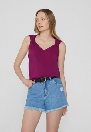 Joven mujer con el pelo largo que lleva una camiseta sin mangas morada metida en unos shorts vaqueros azules de talle alto con un cinturón negro.