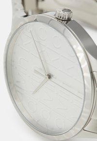 Silver metal watch with a glossy finish and a white dial featuring embossed "Calvin Klein" branding. Thin hour and minute hands.