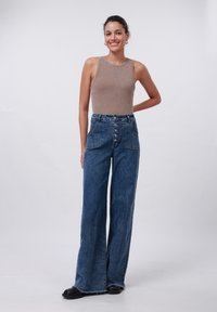 Débardeur beige à côtes associé à un jean en denim bleu à taille haute et jambes larges avec fermeture à boutons et poches avant. Chaussures noires incluses.