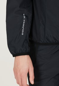 Sort vindjakke med elastiske manchetter, med "ENDURANCE XCS" logo i hvid. Glat tekstur, let materiale og tætsiddende design.