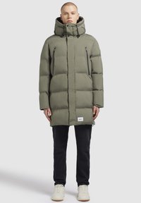 Maslinasto zelena puffer jakna s kapuljačom, s patentnim zatvaračem na grudnim džepovima, blago produženim dizajnom i prednjim etiketam.