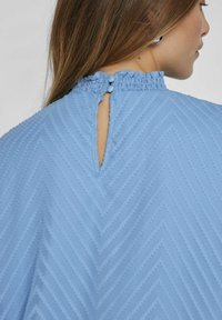Top azzurro chiaro con motivo a chevron testurizzato, collo arricciato elastico e chiusura a occhio di bue sul retro con due bottoni.