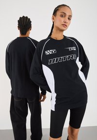 Two people with braided hair wear black and white long-sleeve racing-style shirts with text and graphics, one facing forward, one back.