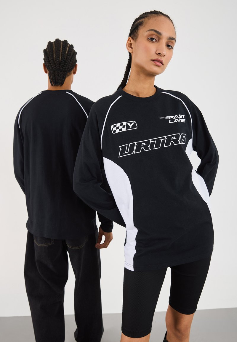 Two people with braided hair wear black and white long-sleeve racing-style shirts with text and graphics, one facing forward, one back.