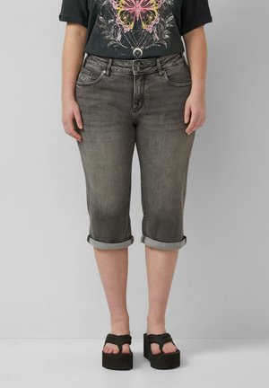 Personne portant un pantacourt en denim gris retroussé, un t-shirt noir avec un motif papillon et des sandales noires à plateforme, debout devant un fond uni.