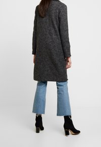 Cappotto lungo grigio scuro realizzato in tessuto tessurizzato, con colletto e maniche attillate. Abbinato a jeans a palazzo azzurri chiari e stivaletti neri alla caviglia.