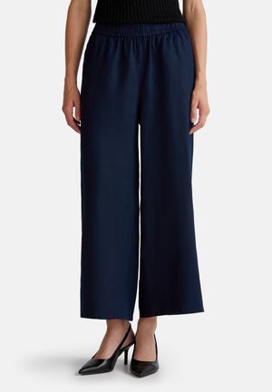 Pantalones de pierna ancha en color marino, hechos de un tejido ligero y con una cinturilla elástica. Combinados con unos tacones altos tipo slingback en color negro.