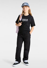Zwarte Vans T-shirt met logo, zwarte broek en zwarte sneakers met witte veters. Lichtblauwe pet en roze geblokte rugzak.