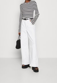 Gestreiftes langärmliges Oberteil in Marineblau und Weiß; kombiniert mit hochtaillierten weißen weiten Hosen. Accessoires sind eine schwarze geflochtene Handtasche und Plateausandalen.