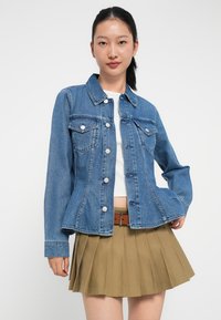 Jeansjakke i mellomblått med klassisk krage, knappedetalj foran, to brystlommer og tilpasset fasong. Båret over en hvit topp og beige plissert skjørt.