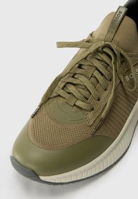 Sapatilha verde oliva com parte superior em tecido de malha, biqueira de borracha e detalhes texturizados. Apresenta atacadores planos e um logotipo circular na lateral.