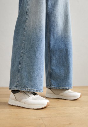 Hvite og beige joggesko med en strukturert overdelflate og en lys farge på sålen, sammen med vide denim-jeans i blått, plassert på et tregulv.