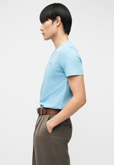 Άνδρας με ανοιχτό μπλε κοντομάνικο πουκάμισο, καφέ ζώνη και γκρι παντελόνι στέκεται προφίλ με το ένα χέρι στην τσέπη, μπροστά σε μονόχρωμο φόντο.