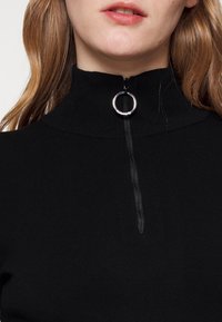 Nahaufnahme eines schwarzen hochgeschlossenes Pullovers mit einem silbernen Reißverschlussgriff am Kragen, getragen von einer Person mit heller Haut und roten Haaren.