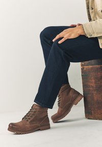 Hnedé kožené členkové čižmy s okrúhlymi špičkami, predným šnurovaním, kontrastným prešívaním a textúrovanými gumovými podrážkami, nosené s tmavými džínami.