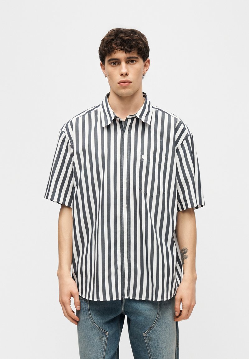 Giovane uomo che indossa una maglietta a maniche corte con righe verticali bianche e nere e jeans blu, in piedi contro uno sfondo chiaro uniforme.