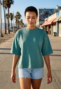 Jeune femme aux cheveux courts et bouclés, portant un T-shirt bleu sarcelle surdimensionné et un short en jean, marchant le long d'une promenade en bord de mer baignée de soleil.
