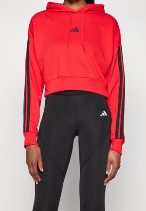 Kobieta ubrana w czerwoną, krótką bluzę z kapturem marki Adidas z czarnymi paskami na rękawach oraz czarne legginsy Adidas z logo na udzie.