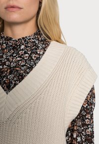 Béžová pletená vesta se širokými žebrovanými rameny, vrstvená přes černou květovanou blůzu s volánkovým výstřihem a knoflíky.