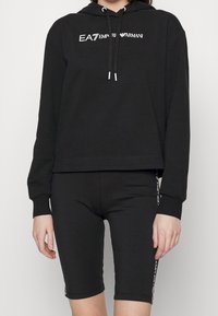 Svart croppad huva med snoddar och EA7-logotyp, kombinerad med svarta cykelshorts som har logotypdetaljer på sidan. Slät, elastisk tyg.