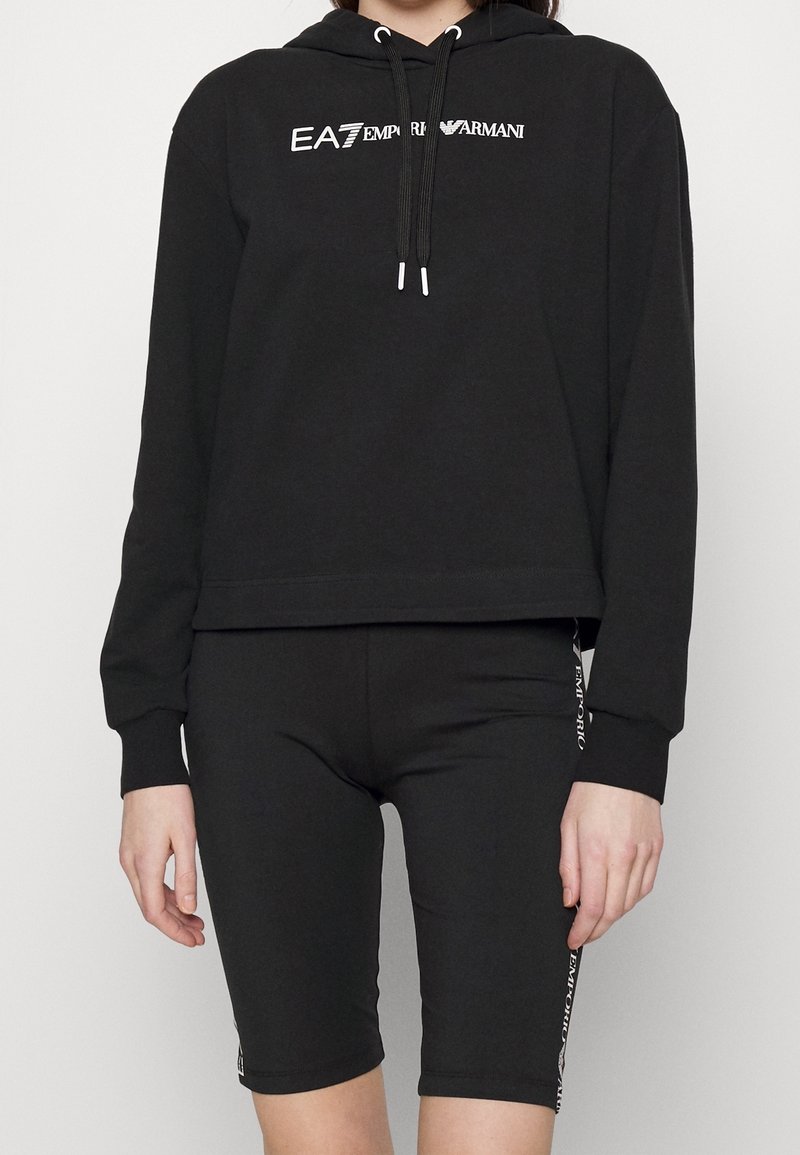 Svart croppad huva med snoddar och EA7-logotyp, kombinerad med svarta cykelshorts som har logotypdetaljer på sidan. Slät, elastisk tyg.