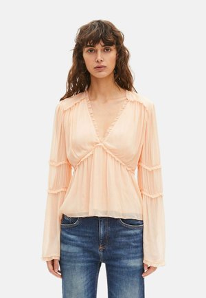 Kobieta ubrana w przezroczystą brzoskwiniową bluzkę z falbaną i długimi rękawami, w połączeniu z niebieskimi dżinsami, stojąca na jednolitym tle.