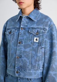 Veste en denim avec un motif camouflage bleu et des nuances claires et foncées. Comprend deux poches poitrine, des boutons en métal et un écusson de marque.