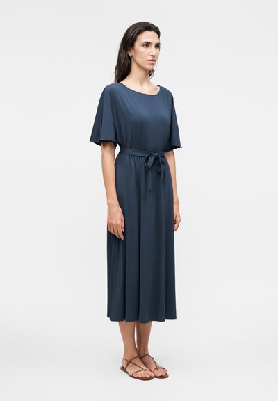 Femme debout portant une robe midi bleu marine à manches courtes avec une ceinture nouée à la taille, associée à des sandales plates marron.
