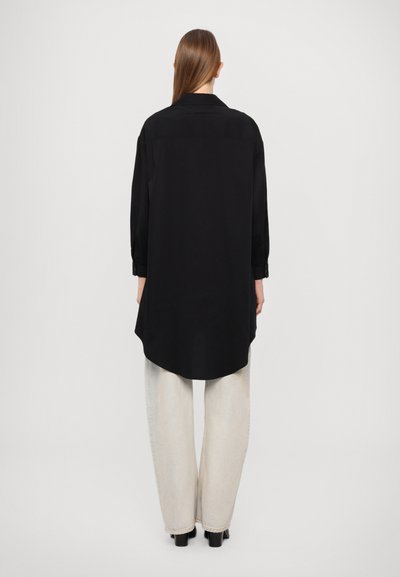 Schwarzes Langarmshirt mit abgerundetem Saum und geknöpften Manschetten, aus glattem Stoff, kombiniert mit hellen, weiten Hosen.