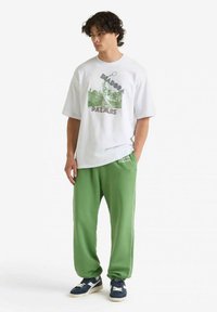 T-shirt bianca in cotone con design grafico verde e testo, abbinata a pantaloni della tuta verdi con polsini elasticizzati e strisce laterali a contrasto.