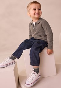 Grauer Strickpullover mit Reißverschluss am Kragen, blaue Jeanshose und weiße Sneakers mit roten Akzenten, sitzend auf weißen Blöcken vor beigem Hintergrund.