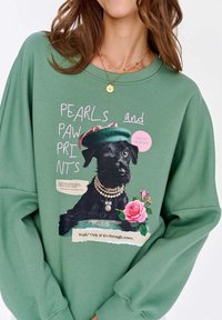 Zielona bluza z grafiką czarnego psa w zielonej czapce i perełkach, ozdobiona różowymi różami i zabawnym tekstem.