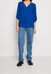Blauwe, textuurrijke top met een v-hals en opgerolde mouwen, gecombineerd met gestreepte blauwe broeken met geometrische vormen. Zwarte schoenen maken de outfit compleet.