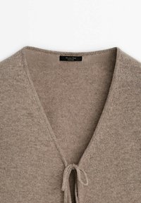 Beige neulepusero V-kaula-aukkolla ja solmittavalla kiinnityksellä, jossa on pehmeä pinta ja yksinkertainen muotoilu. Etiketissä lukee "Massimo Dutti, Made in China."