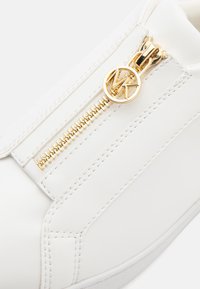 Baskets blanches avec une texture en cuir lisse, dotées de zips en métal doré et d'un logo en métal accentué. Coutures détaillées le long des côtés.