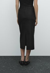 Person som står med ryggen mot betraktaren, iklädd en svart midikjol med en slits bak och svarta platta skor på ett mörkt fläckigt golv.