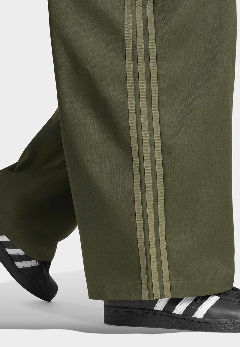 Pantalon vert olive avec trois bandes verticales sur les côtés, associé à des baskets noires avec des bandes blanches sur les côtés.