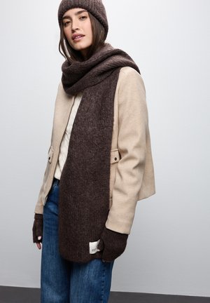 Veste en laine beige sur un pull crème, associée à une écharpe en tricot marron foncé et des gants sans doigts. Un jean bleu complète la tenue.