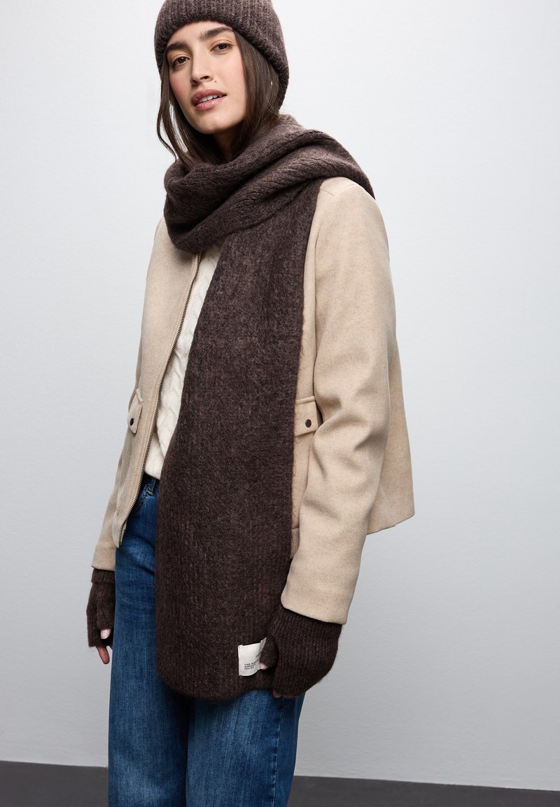 Veste en laine beige sur un pull crème, associée à une écharpe en tricot marron foncé et des gants sans doigts. Un jean bleu complète la tenue.