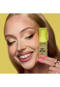 Femme souriante avec un fard à paupières jaune et des ongles roses tenant le produit de maquillage NYX Plump Right Back près de son visage sur un fond jaune.