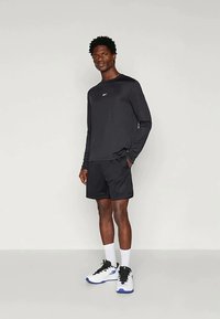 Άνδρας που στέκεται φορώντας μαύρο πουκάμισο με μακριά μανίκια, μαύρες βερμούδες, λευκές κάλτσες και λευκά αθλητικά παπούτσια με μπλε σόλες, μπροστά σε λείο φόντο.