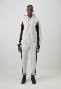 Sudadera con cremallera de color gris claro con paneles laterales de color gris oscuro, combinada con joggers de gris claro a juego. Tejido suave, corte relajado y capucha ajustable.
