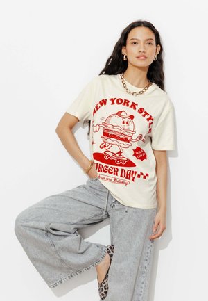 Femme portant un T-shirt blanc avec un hamburger de dessin animé rouge, sur un skateboard et un jean large gris, posant avec une jambe levée.