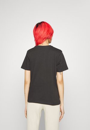 Person med lyst rødt hår iført en simpel sort t-shirt og hvide bukser, stående med ryggen til mod en lys grå baggrund.