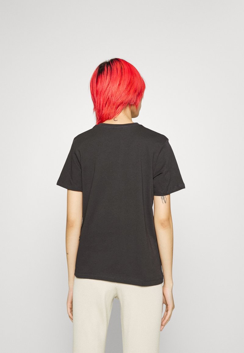 Personne aux cheveux rouge vif portant un t-shirt noir uni et un pantalon blanc, debout de dos sur un fond gris clair.