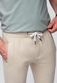 Beige sweatpants met een elastische tailleband en een koord, voorzien van zijzakken en een gladde textuur, gecombineerd met een lichtgroen shirt.