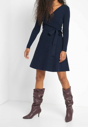 Woman wearing a navy blue long-sleeve wrap dress with a waist tie, paired with slouchy dark brown leather knee-high boots with kitten heels.