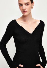 Schwarzes, geripptes Langarmoberteil mit tiefem V-Ausschnitt, figurbetonter Silhouette und ausladenden Bündchen. Glatte Textur, minimalistisches Design und einfarbig.