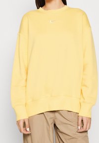 Sudadera amarilla con cuello redondo y puños acanalados. Presenta un pequeño logo blanco de Nike en el pecho. Tejido suave y diseño de corte holgado.